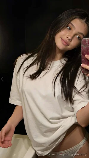 Woman with long dark hair in loose white t-shirt and underwear, taking mirror selfie indoors.