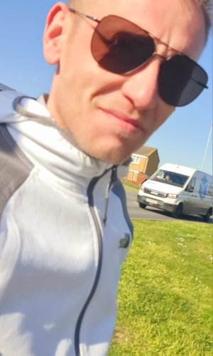 Man in gray hoodie and sunglasses outdoors by road.