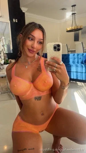 Woman in orange lingerie posing in a modern living room with tattoos visible.