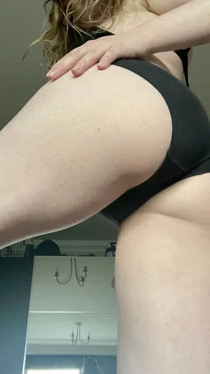 Woman in black underwear posing in bedroom, hand on thigh, blonde hair visible.