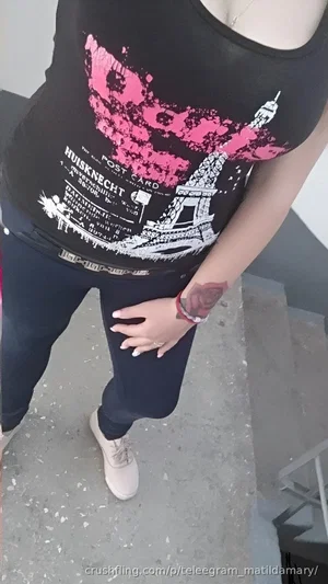 Woman in black tank top with graphic, dark pants, and white sneakers standing indoors.