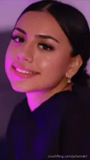 Woman with dark hair and makeup, wearing dark top, smiling in indoor setting with purple lighting.