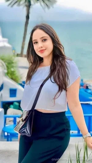 Woman with long dark hair in a white crop top and dark pants, standing outdoors near a beach with