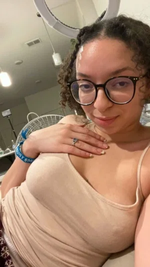 Woman with curly hair and glasses in a beige tank top, posing indoors near a ring light.