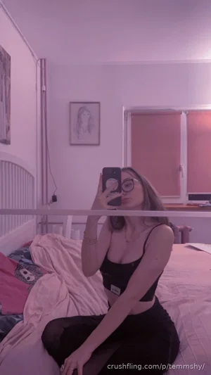 Woman in black crop top and pants sits on bed, taking mirror selfie in bedroom with pink lighting.