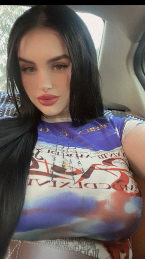 Woman with long black hair in colorful graphic tee, sitting in car.
