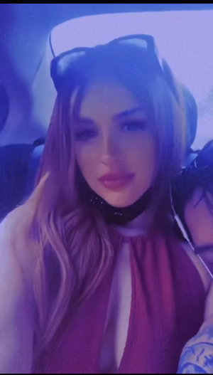 Woman with long blonde hair in a car, wearing a dark top and choker, sunglasses on head.