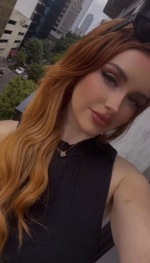 Woman with long red hair in black sleeveless top, taking selfie outdoors with city view.