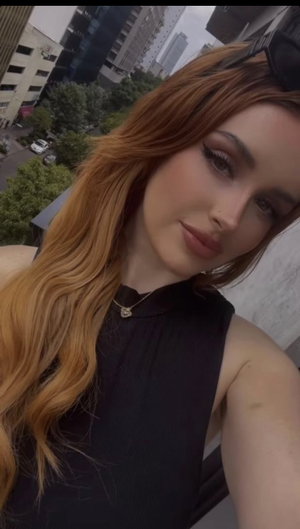 Woman with long red hair in black sleeveless top, taking selfie outdoors with city view.
