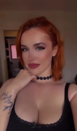 Woman with red hair and black lace top posing indoors, hand on chin.