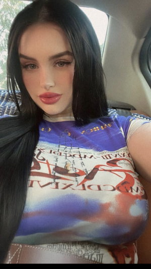 Woman with long black hair wearing colorful graphic top, sitting in car.