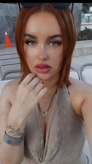 Woman with short red hair in a gray halter top, posing outdoors near chairs.