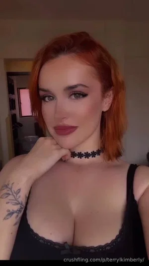 Woman with red hair, black lace top, choker, tattoo on arm, indoors.
