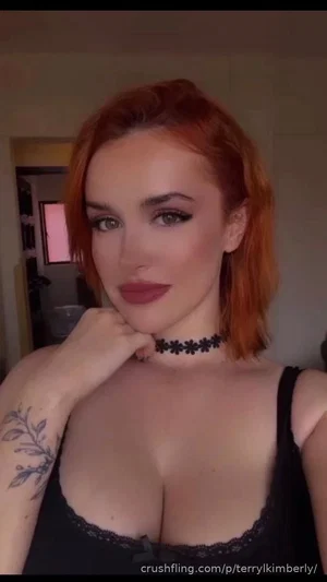 Woman with red hair and tattoos wears black lace top, posing indoors.