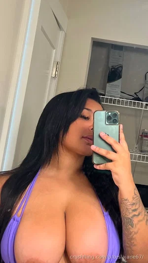 Woman with long black hair in purple bikini top, holding phone for mirror selfie in indoor setting.