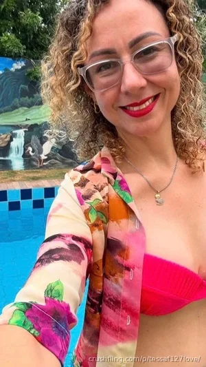 Woman with curly hair and glasses in a pink bikini top and floral shirt by a pool.