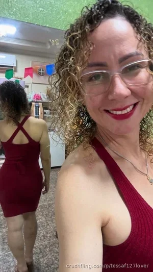 Woman with curly hair and glasses in a red dress, taking a mirror selfie indoors.