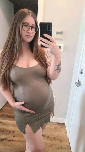 Woman with long brown hair and glasses in olive green dress, holding phone for mirror selfie