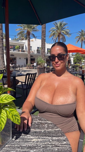 Woman in brown bandeau top sitting at outdoor cafe under umbrella, palm trees in background.