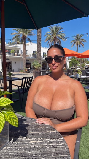 Woman in brown bandeau top sitting at outdoor table under umbrella, palm trees in background.