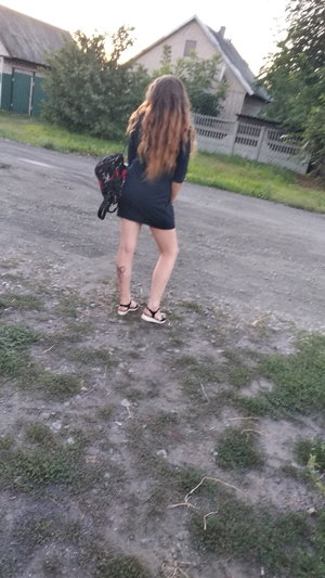 Woman in short black dress and sandals stands outdoors on gravel.