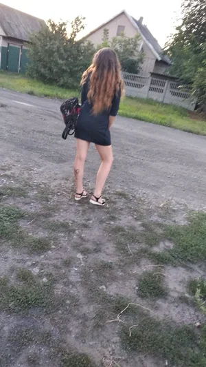 Woman with long hair in black dress standing outdoors on gravel, holding bag.