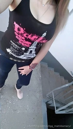Woman in black tank top and jeans standing near stairs indoors.