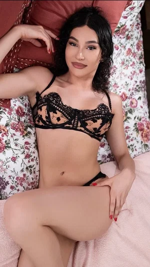 Woman in black lace lingerie posing on floral pillows in a bedroom.