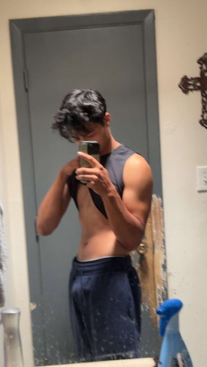 Man with dark hair and muscular build takes mirror selfie in bathroom wearing black tank top and