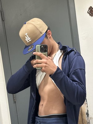 Man in blue hoodie and cap takes mirror selfie, showing bare torso and abs.