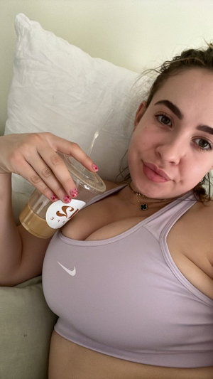 Curvy woman in lavender sports bra holding drink on bed.