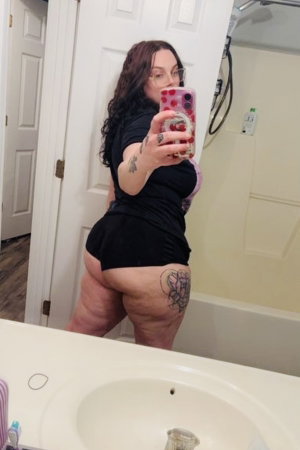 Curvy woman with glasses taking bathroom mirror selfie in black outfit.