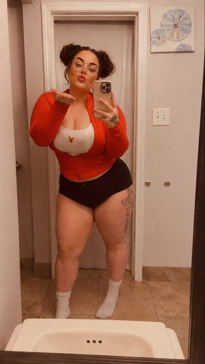 Woman in red cardigan, white bra, black shorts, white socks, taking mirror selfie in bathroom.