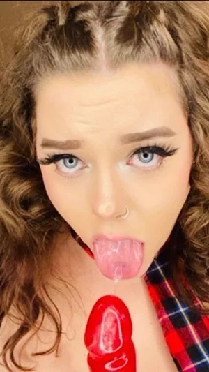 Woman with curly hair and makeup sticks out tongue, holding red object, wearing plaid shirt.