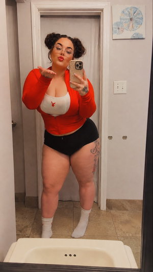 Woman in red cardigan, white bra, black shorts, white socks, taking mirror selfie in bathroom.