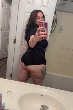 Woman with curly hair taking mirror selfie in bathroom wearing black outfit.