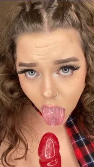 Woman with curly hair and makeup, wearing plaid shirt, holding red object near mouth.