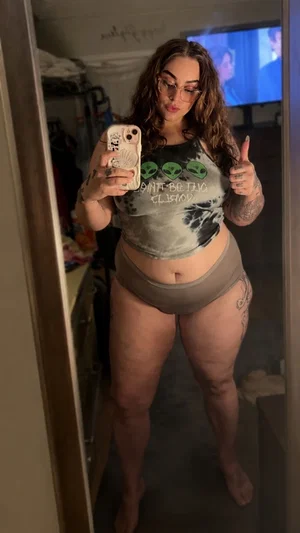 Woman with curly hair and tattoos wears crop top and underwear, posing in mirror selfie indoors.