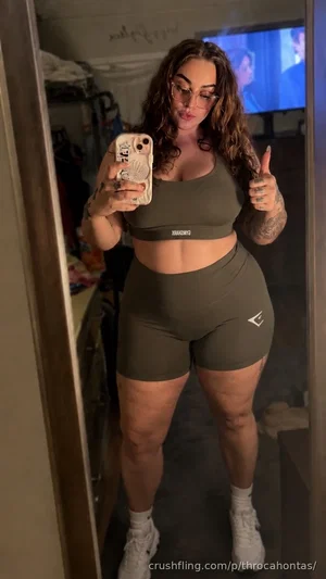 Woman with curly hair and tattoos wears olive green sports bra and shorts, taking mirror selfie in
