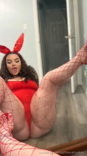 Woman in red bodysuit and fishnet stockings, wearing devil ears, posing on floor indoors.