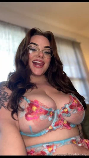 Woman with long brown hair and glasses wearing floral lace lingerie, smiling in a bedroom.