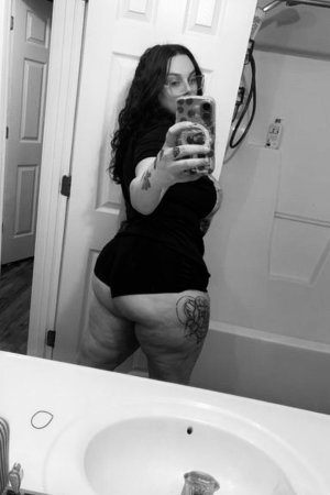 Woman with tattoos in black top and shorts taking mirror selfie in bathroom.