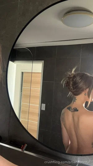 Woman with tattoos on back, hair in bun, seen from behind in mirror selfie in bathroom.