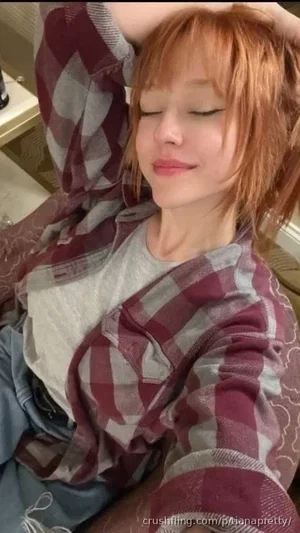 Woman with short red hair in plaid shirt and gray tee, sitting indoors with eyes closed.