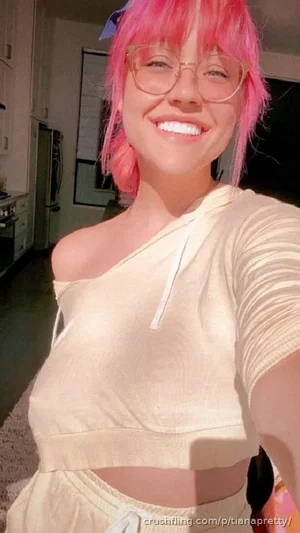 Woman with pink hair and glasses in a beige one-shoulder crop top, smiling in a kitchen.