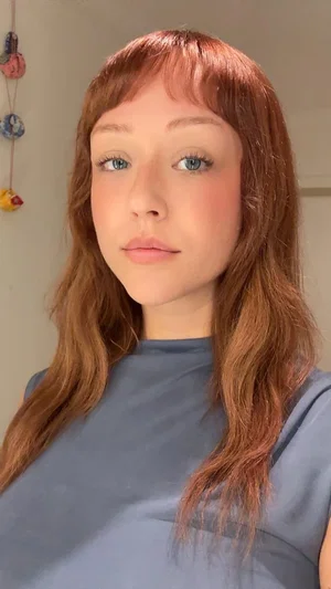 Woman with red hair and blue eyes wearing a gray top indoors.