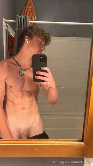 Shirtless man with curly hair taking mirror selfie in bathroom, wearing black shorts and necklace.