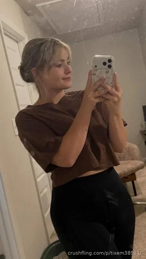 Woman with blonde hair in a brown crop top and black leggings taking a mirror selfie indoors.
