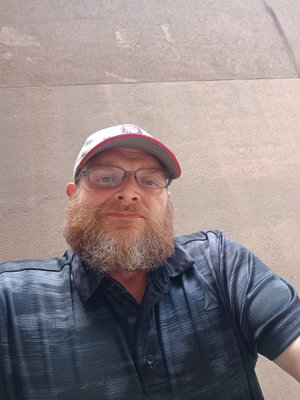 Man with red beard and glasses wearing cap and polo shirt outdoors.