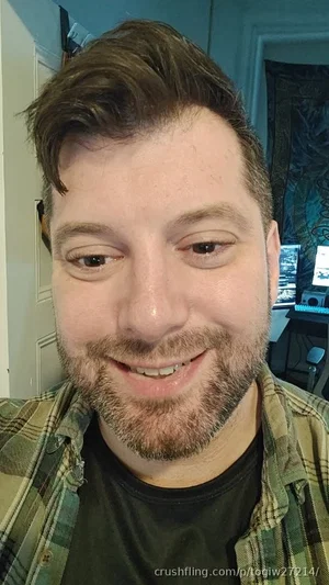 Man with short brown hair and beard, wearing plaid shirt over black tee, indoors.